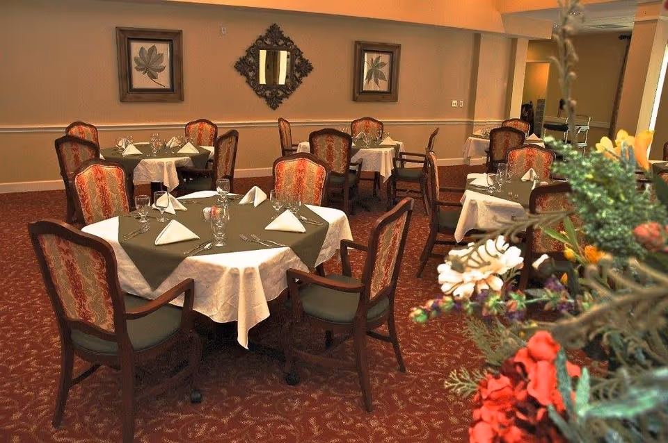 A dining room with several round tables covered with white tablecloths and green overlays, each set with folded napkins, glasses, and silverware. The chairs have wooden frames with patterned upholstery. The room has framed leaf artwork and a decorative mirror on the wall, with a floral arrangement visible in the foreground.