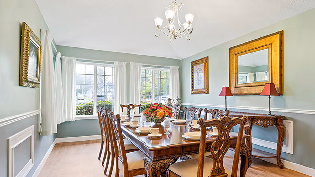 A well-lit dining room with a long wooden dining table set with plates, cups, and glasses. The room features large windows with white curtains, light green walls, a chandelier, framed artwork, a large gold-framed mirror, and a wooden side table with two red lamps.