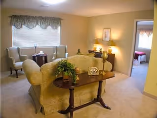 A cozy living room area with a beige upholstered sofa facing away from the camera, a wooden console table behind it with decorative items including a plant and a framed photo. Two armchairs and a small round table are positioned near a window with blinds and a valance. A sideboard with lamps and framed artwork is against the far wall. An open doorway leads to another room with a bed visible.