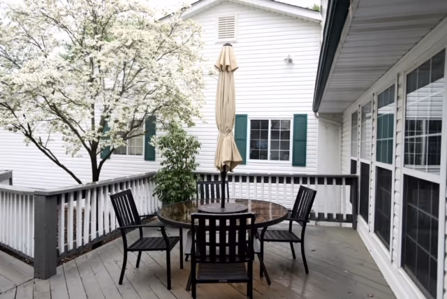 Outdoor patio area with a round table and four black chairs. A closed beige umbrella is in the center of the table. The patio is surrounded by a white railing and adjacent to a white building with green window shutters. A tree with white blossoms is visible on the left side.