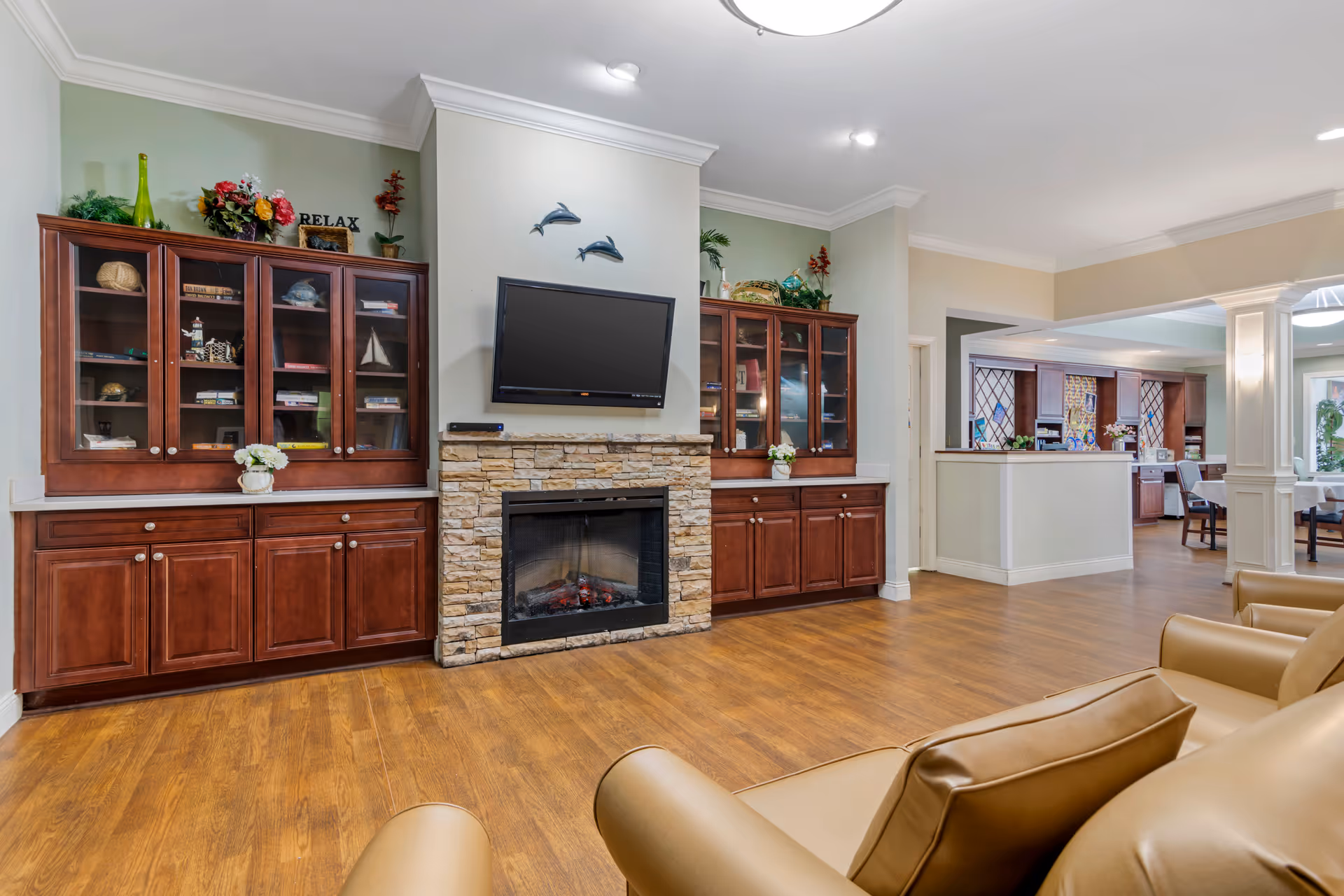 A bright community living room with built-in wooden cabinets flanking a stone fireplace and wall-mounted TV, leather chairs and an open dining area visible behind.
