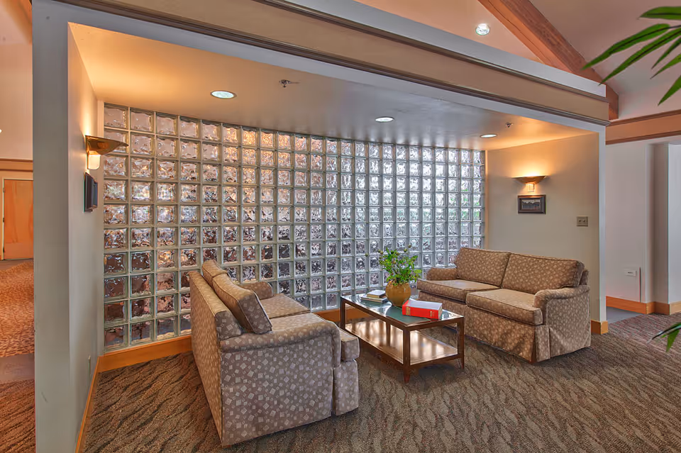 A cozy seating area in a senior living facility with two patterned sofas facing each other across a wooden coffee table. The table holds a potted plant and some books. The background features a large glass block wall allowing natural light to filter in. The room has carpeted flooring, warm wall sconces, and exposed wooden ceiling beams.