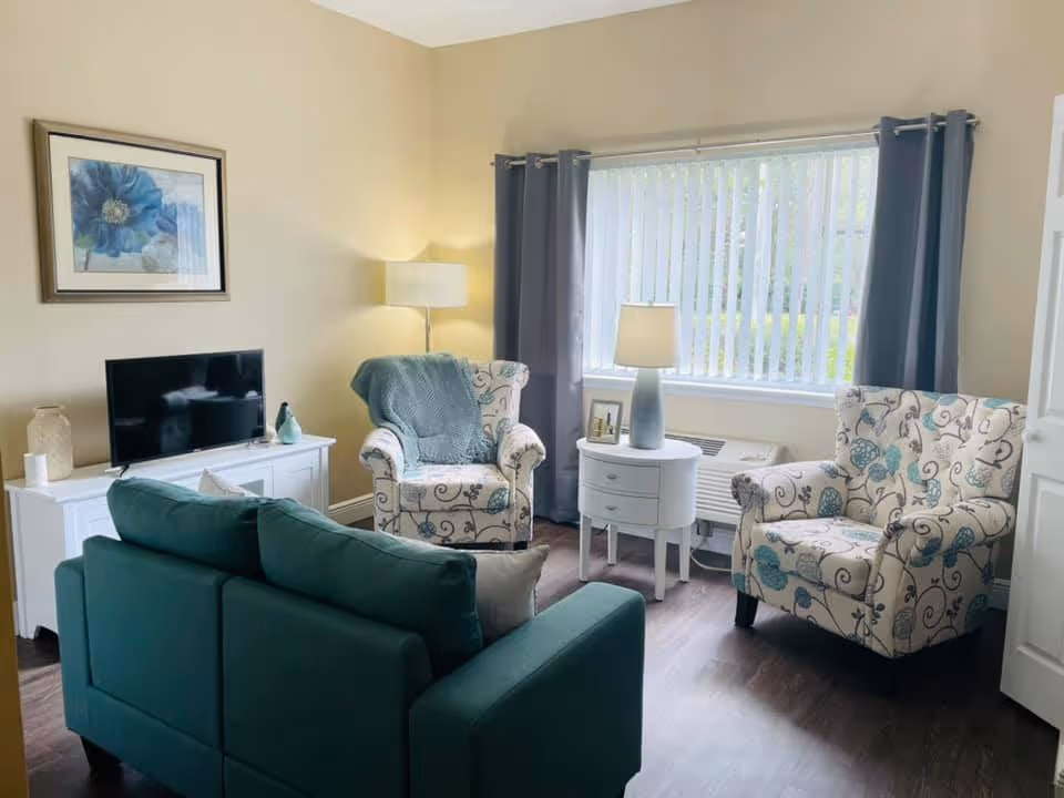 A cozy living room with a teal sofa facing two patterned armchairs. There is a white TV stand with a flat-screen TV, a floor lamp, and a small round white side table with a lamp and a framed photo. A large window with vertical blinds and gray curtains lets in natural light.