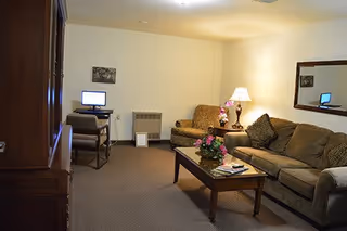A cozy living room with a brown sofa, a matching loveseat, a wooden coffee table with flowers and magazines, a table lamp, a large mirror on the wall, and a small desk with a computer and chair in the corner.