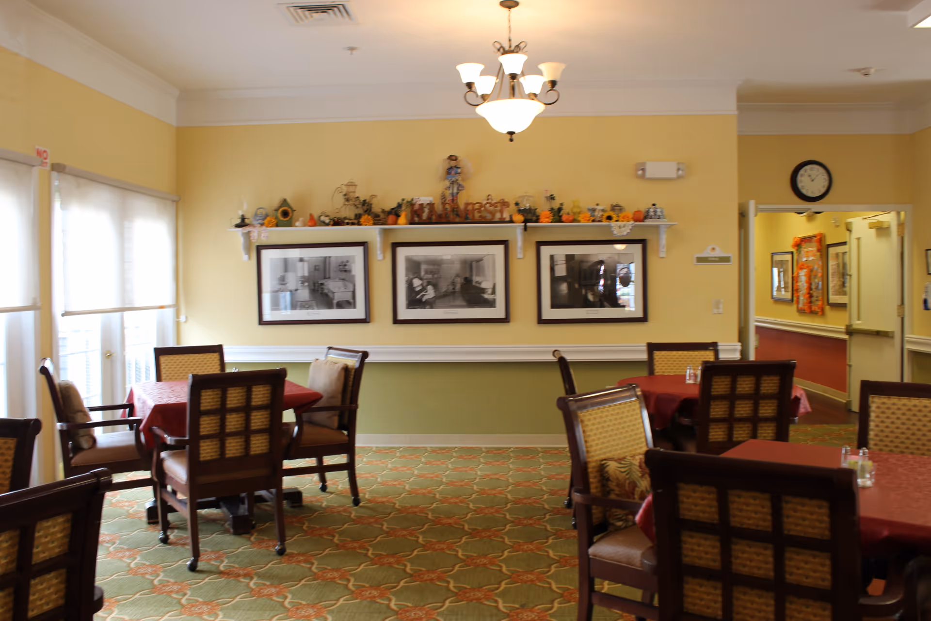 Dining room with several tables and chairs, framed black-and-white photos on a yellow wall and a decorative shelf above.