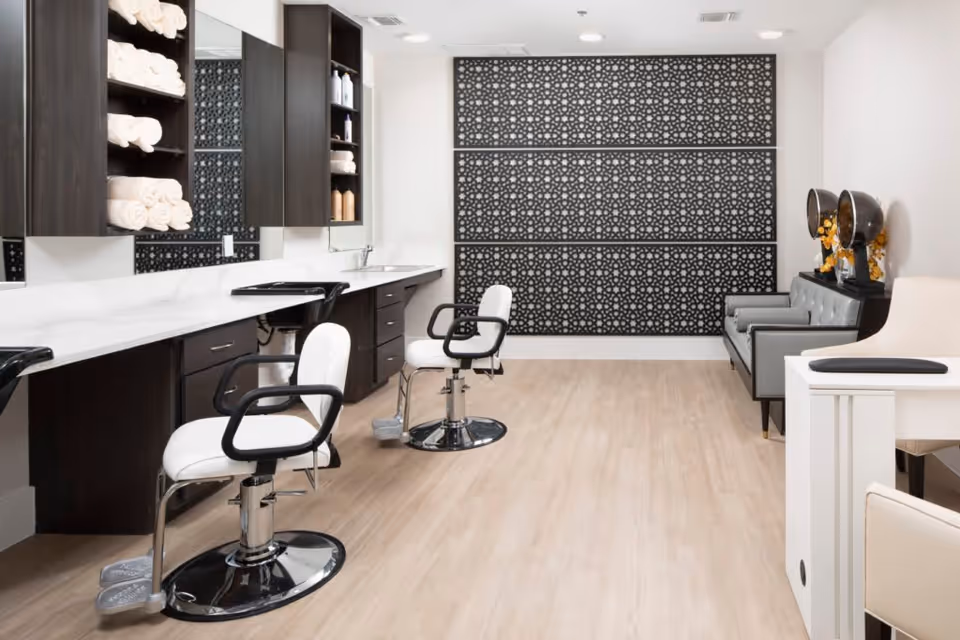 Well-lit salon interior with styling chairs, a long counter with mirrors and shelves of towels, and a seating area against a decorative wall.