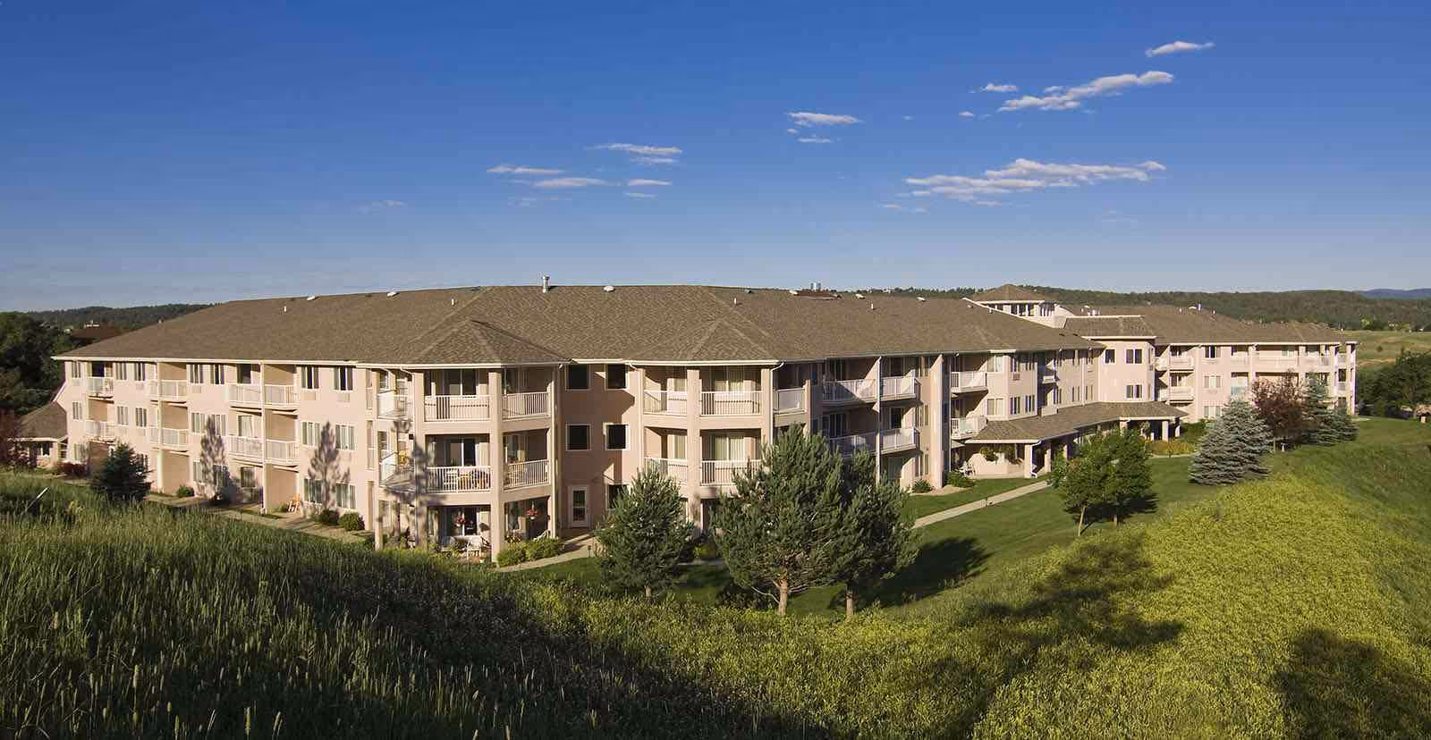Three-story beige senior living building with balconies on a grassy hillside under a blue sky.