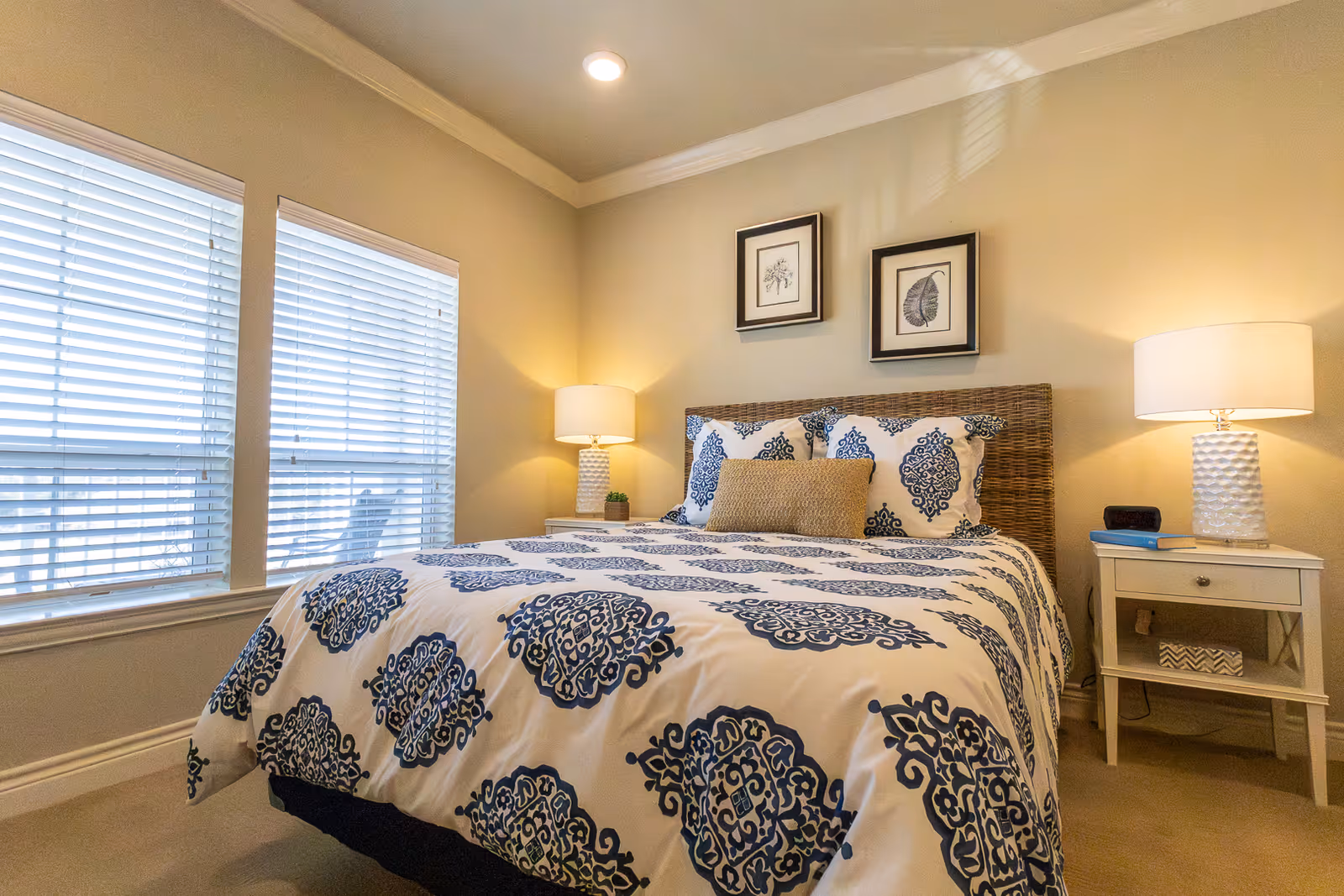A cozy bedroom with a bed featuring a white and blue patterned comforter and pillows. The bed has a woven headboard and is flanked by two white nightstands, each with a lamp. Two framed botanical prints hang on the wall above the bed. There are two large windows with white blinds on the left side of the room.