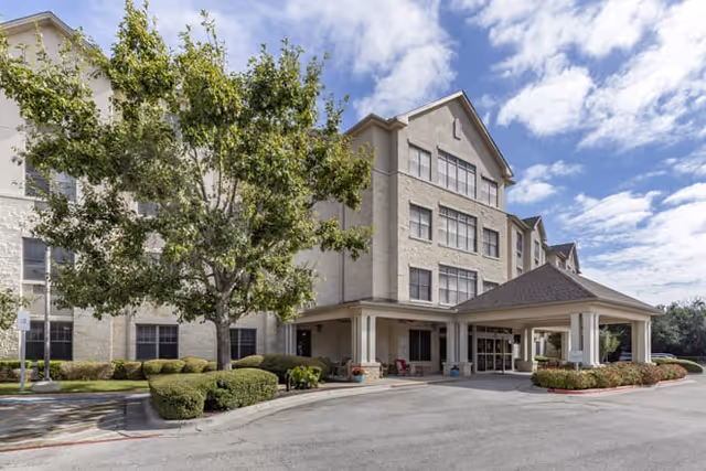 Exterior view of a multi-story senior living facility building with beige brick walls, multiple windows, a covered entrance with columns, landscaped bushes, and a large tree in front under a partly cloudy sky.