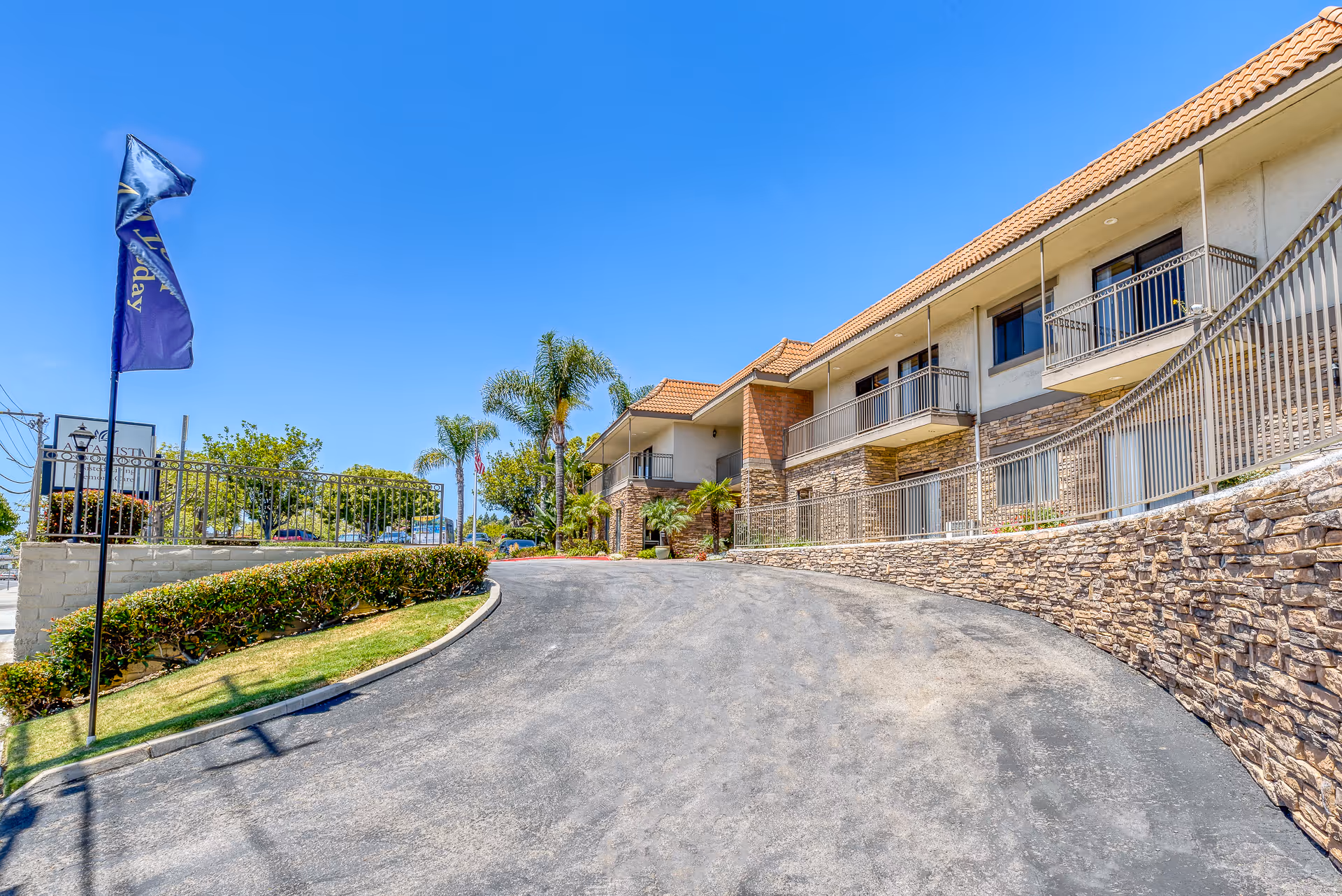 Exterior view of Alta Vista Senior Living building with a curved driveway, stone walls, balconies, palm trees, and a clear blue sky.