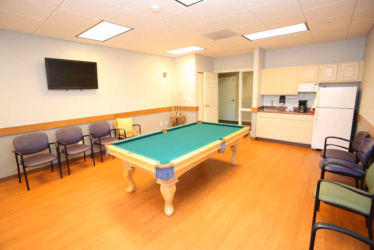 A bright common room with a pool table in the center, chairs along the walls, a wall-mounted TV, and a small kitchenette.