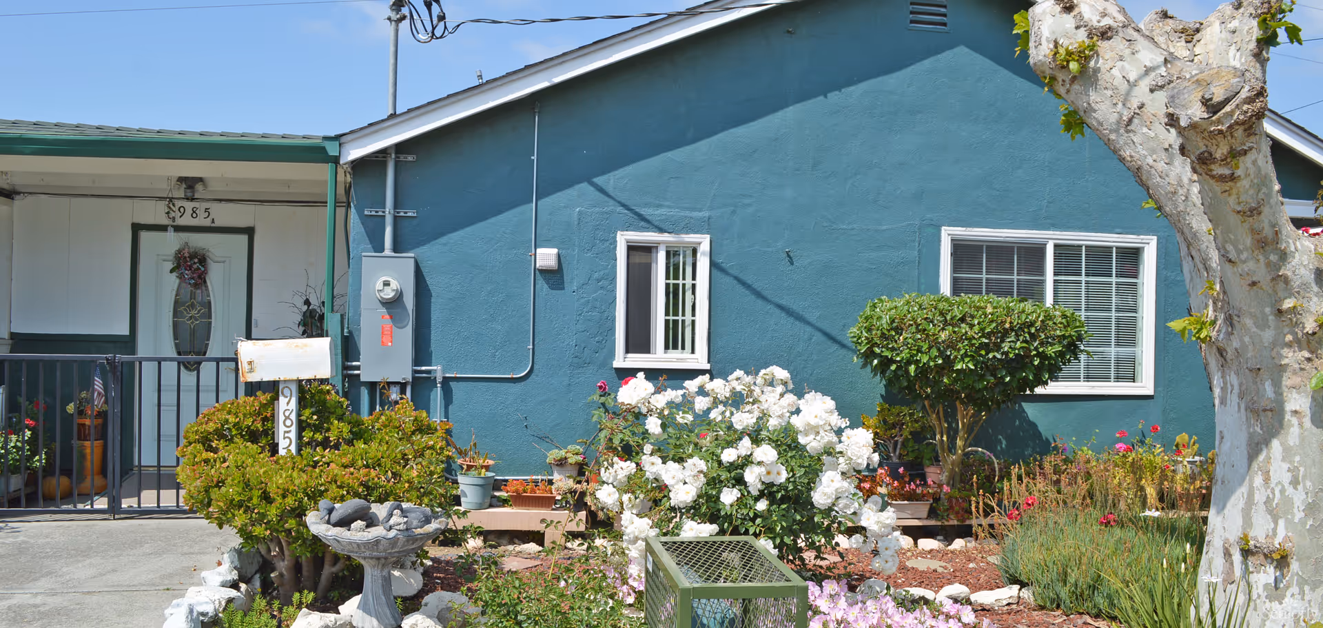 Front view of a single-story teal house with a porch entrance, mailbox, and a flower-filled front yard.