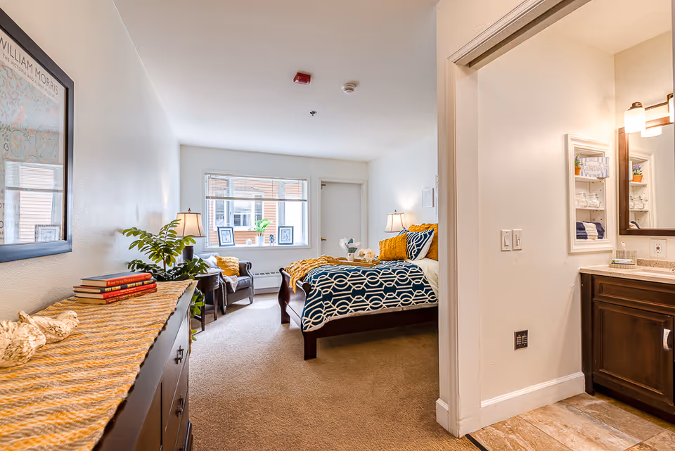 Bright furnished bedroom suite with a bed, nightstands, a seating area by the window, and a view into the bathroom vanity.