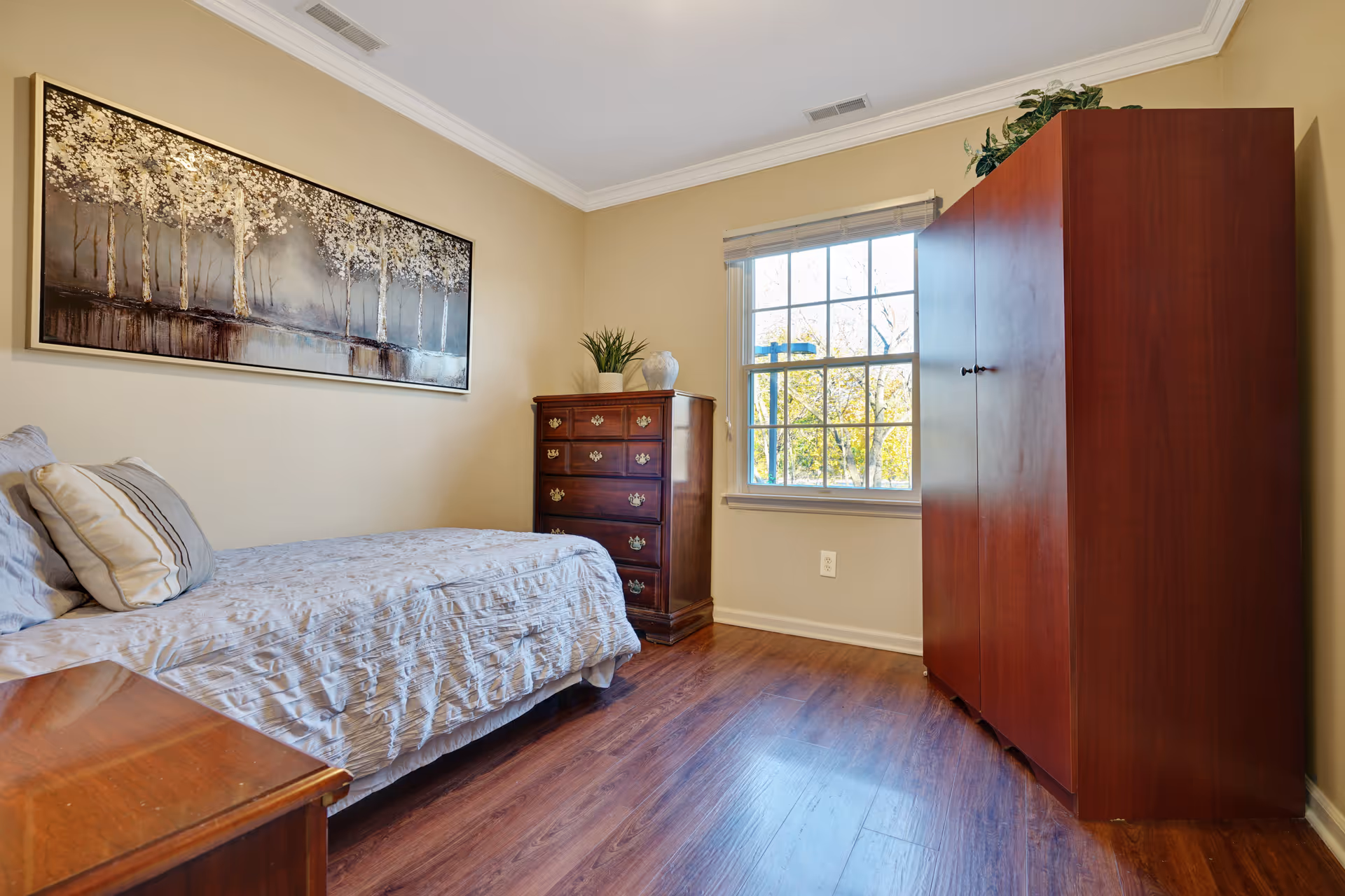 A cozy bedroom with a single bed covered in a light-colored bedspread and pillows. There is a wooden dresser with drawers and decorative items on top, a large wooden wardrobe, and a window letting in natural light. A framed artwork of trees hangs on the wall above the bed. The room has wooden flooring and beige walls.