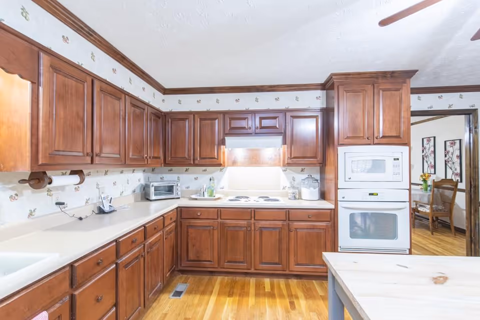 A bright kitchen with wooden cabinets and drawers, a white countertop, a built-in microwave and oven, an electric stovetop with a range hood, a toaster oven, and a paper towel holder. The kitchen has hardwood flooring and floral wallpaper. Through an open doorway, a dining area with a wooden table and chairs and floral wall art is visible.