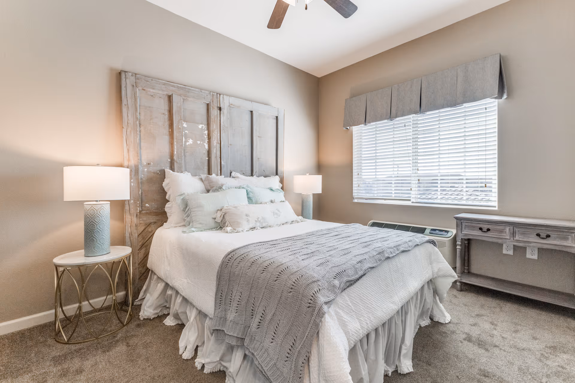 A cozy bedroom with a bed featuring a rustic wooden headboard, white bedding, multiple pillows, and a gray knitted throw blanket. There are two bedside tables with matching lamps on either side of the bed. A window with white blinds and a gray valance lets in natural light. A small wooden desk is positioned against the wall under the window, and the room has beige walls and carpeted flooring.