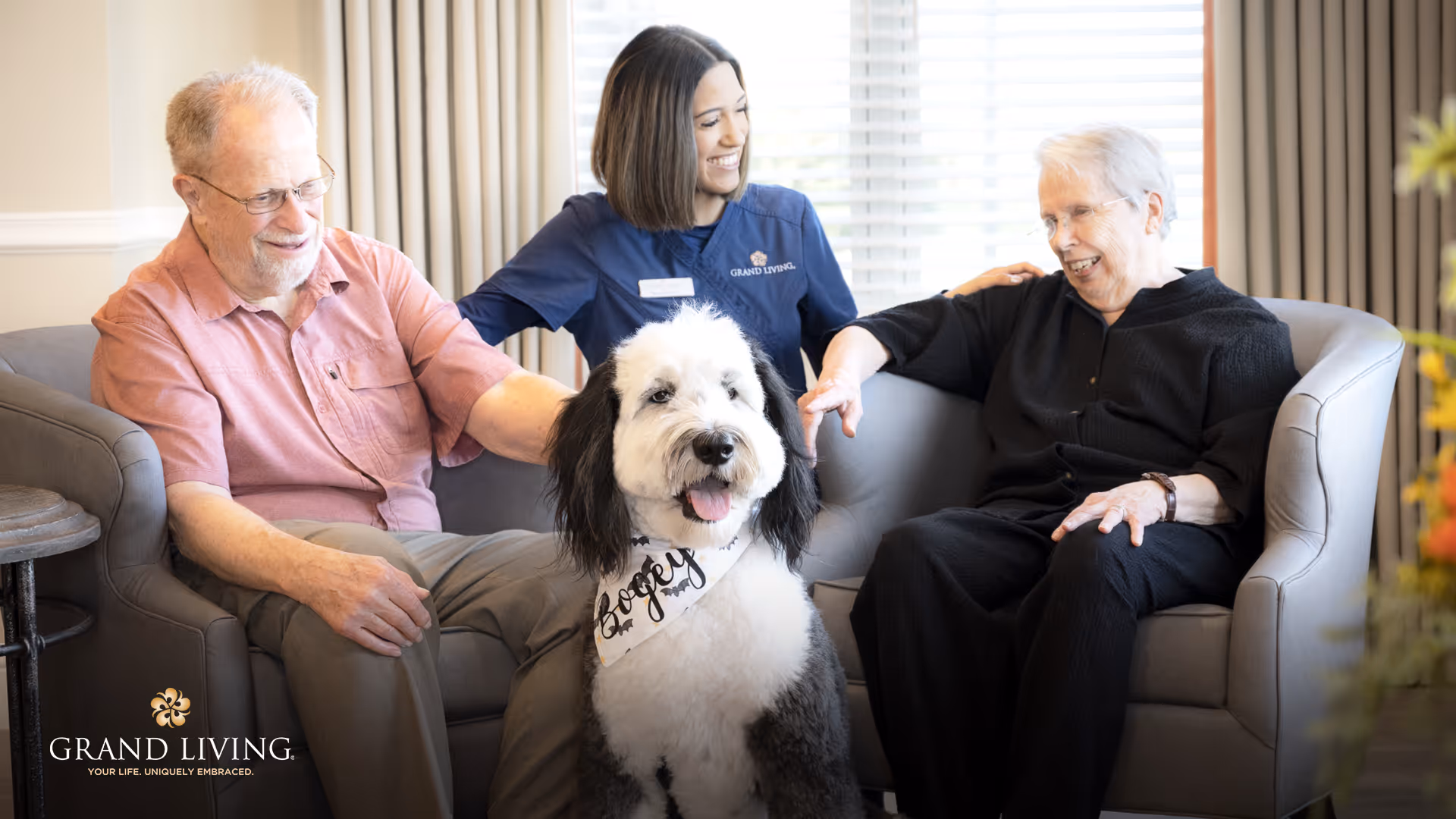 Two elderly individuals sitting on a couch with a smiling caregiver in a navy blue uniform behind them. A large fluffy black and white dog wearing a bandana with the name 'Bogey' sits in front of them. The setting appears to be a bright, comfortable living room with a window and curtains in the background.