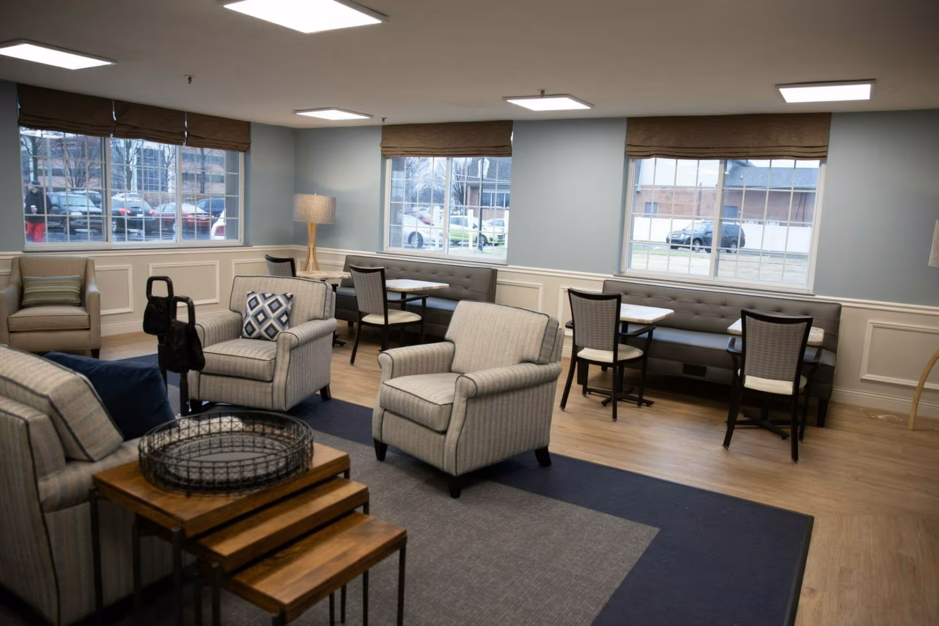 A well-lit common area with comfortable armchairs, a sofa, and small tables. There are three large windows with brown blinds, letting in natural light and showing a view of parked cars outside. The room has light blue walls with white wainscoting and wooden flooring, creating a cozy and inviting atmosphere.