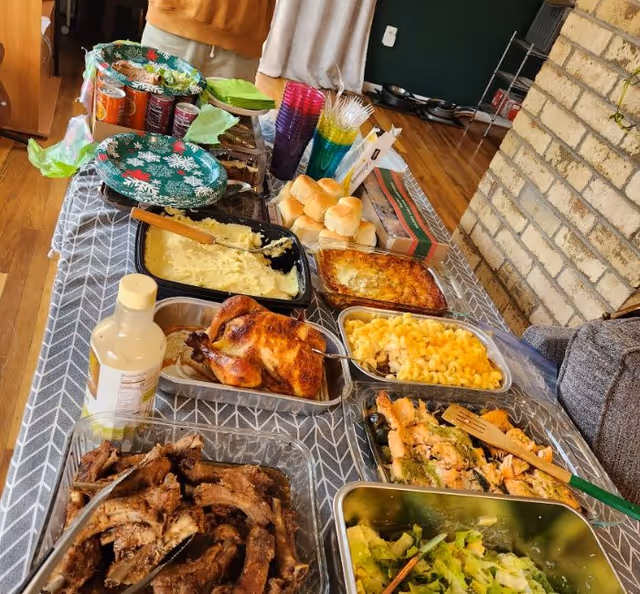 A buffet-style table set with various dishes including roasted chicken, ribs, mashed potatoes, macaroni and cheese, salad, dinner rolls, and a casserole. There are colorful plastic cups, canned drinks, and utensils on the table. The setting appears to be indoors with a brick wall and wooden floor visible.