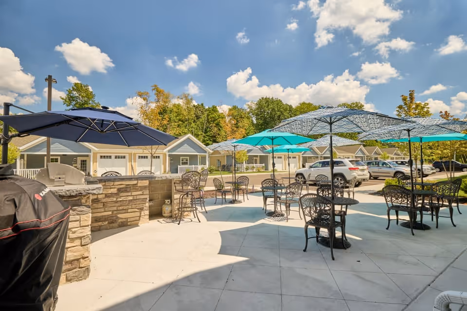 Outdoor patio with metal tables and chairs under blue and teal umbrellas in front of a row of cottages and parked cars.
