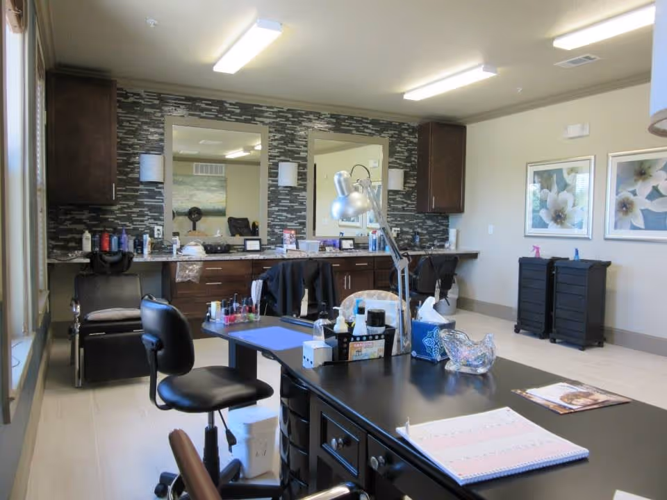 Interior view of a salon or beauty care room with a black manicure table in the foreground, equipped with nail polish bottles, a desk lamp, and other manicure tools. The background features a wall with two large mirrors, dark wood cabinets, and shelves holding various hair and beauty products. Two black chairs are positioned in front of the mirrors, and two framed floral artworks hang on the adjacent wall.