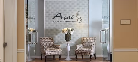 Two patterned armchairs with wooden legs are positioned on either side of a small white pedestal table holding a vase of white flowers. Behind them is a wall with a mosaic tile design and the words 'Açaí BEAUTY IN ITS RAREST FORM' displayed. The area is framed by glass doors with metal handles, and a beige wall with a sign labeled 'SPA' is visible on the right side.