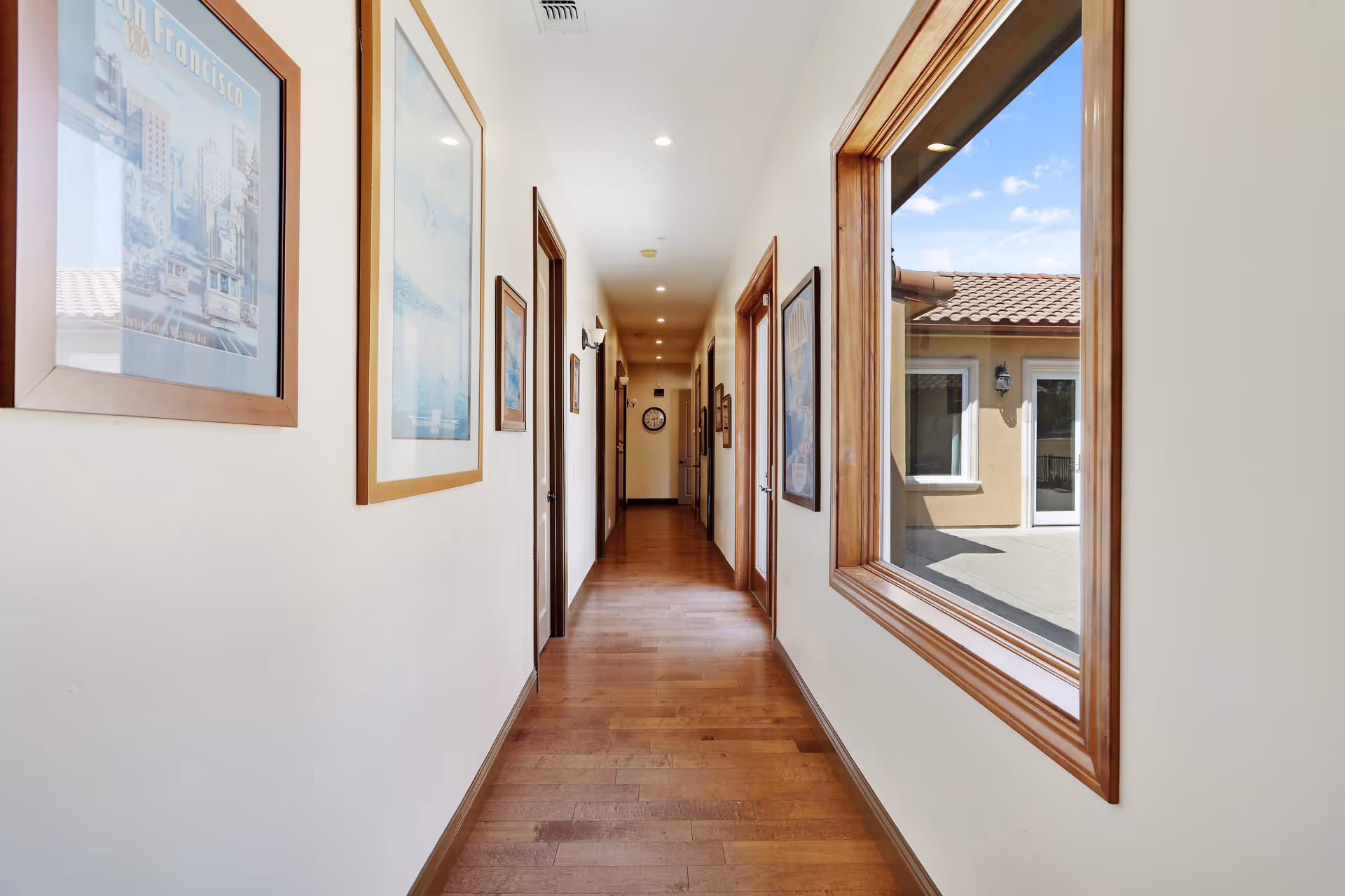 A long, narrow hallway with wooden flooring and cream-colored walls. Several framed pictures hang on the walls, and there are multiple doorways along the corridor. On the right side, a large window reveals an outdoor courtyard with a tiled roof and a clear blue sky. The hallway is well-lit with recessed ceiling lights.
