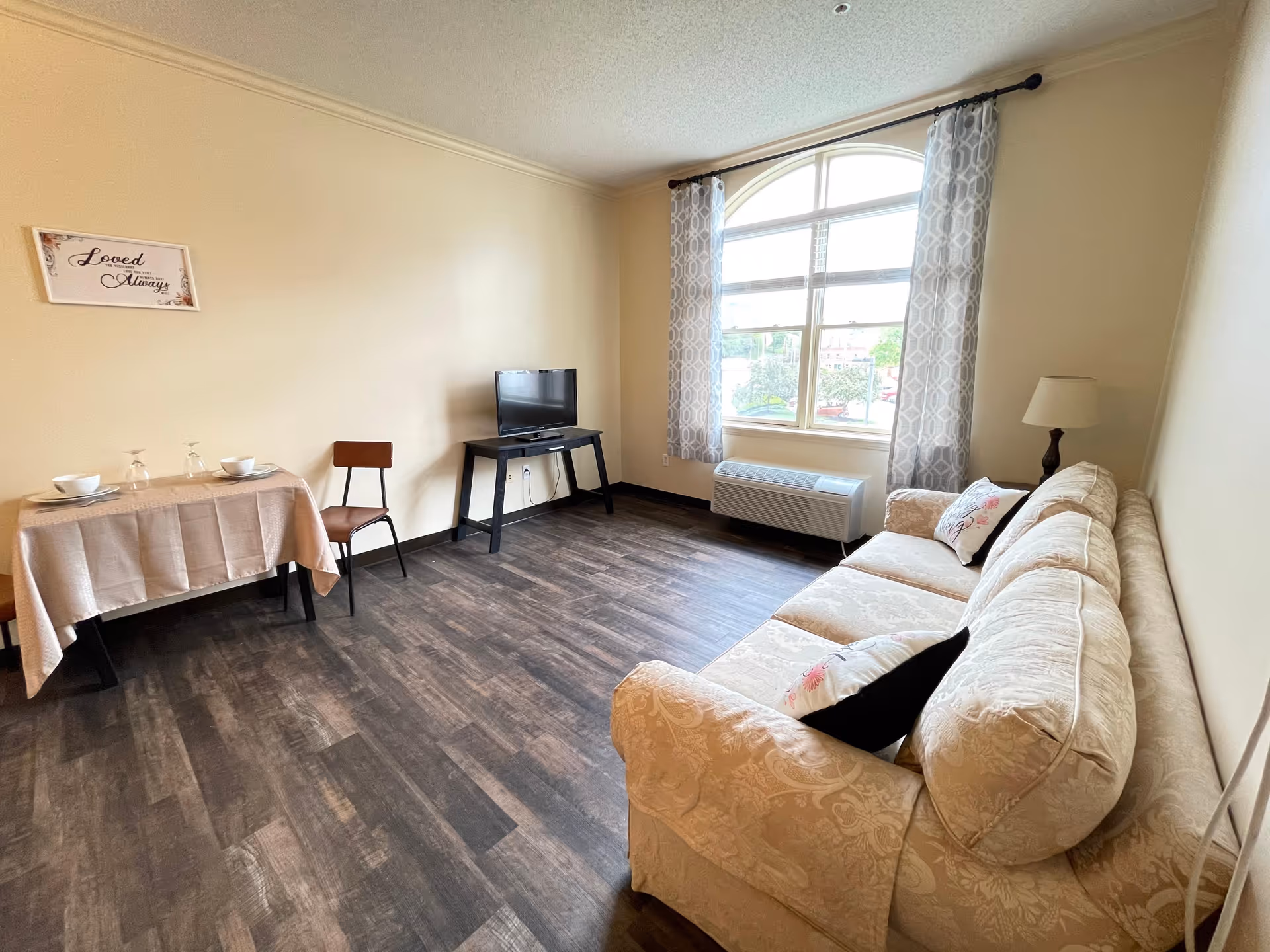 A cozy living room with a beige patterned sofa adorned with decorative pillows, a small table with a TV on it, a dining table set for two with a tablecloth, and a large window with patterned curtains letting in natural light. A floor lamp is positioned next to the sofa, and a wall decoration reads 'Loved Always'.