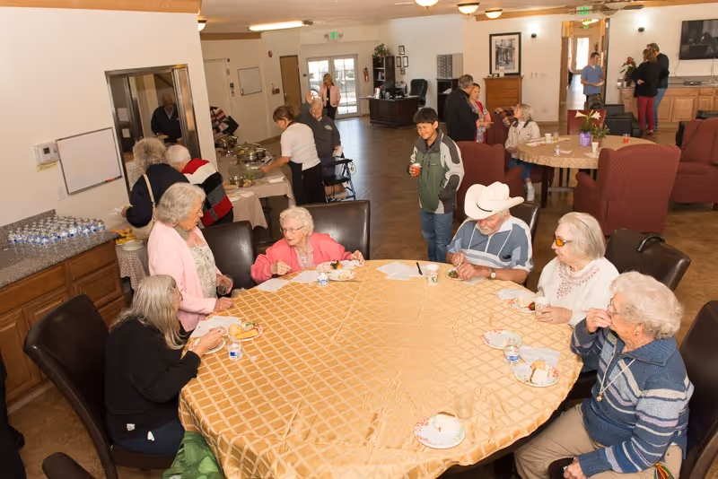 A group of elderly people sitting around a large round table covered with a beige tablecloth, eating and drinking in a communal dining area. Other residents and staff are seen in the background near a serving counter and sitting at another table. The room is well-lit with overhead lights and has a warm, welcoming atmosphere.