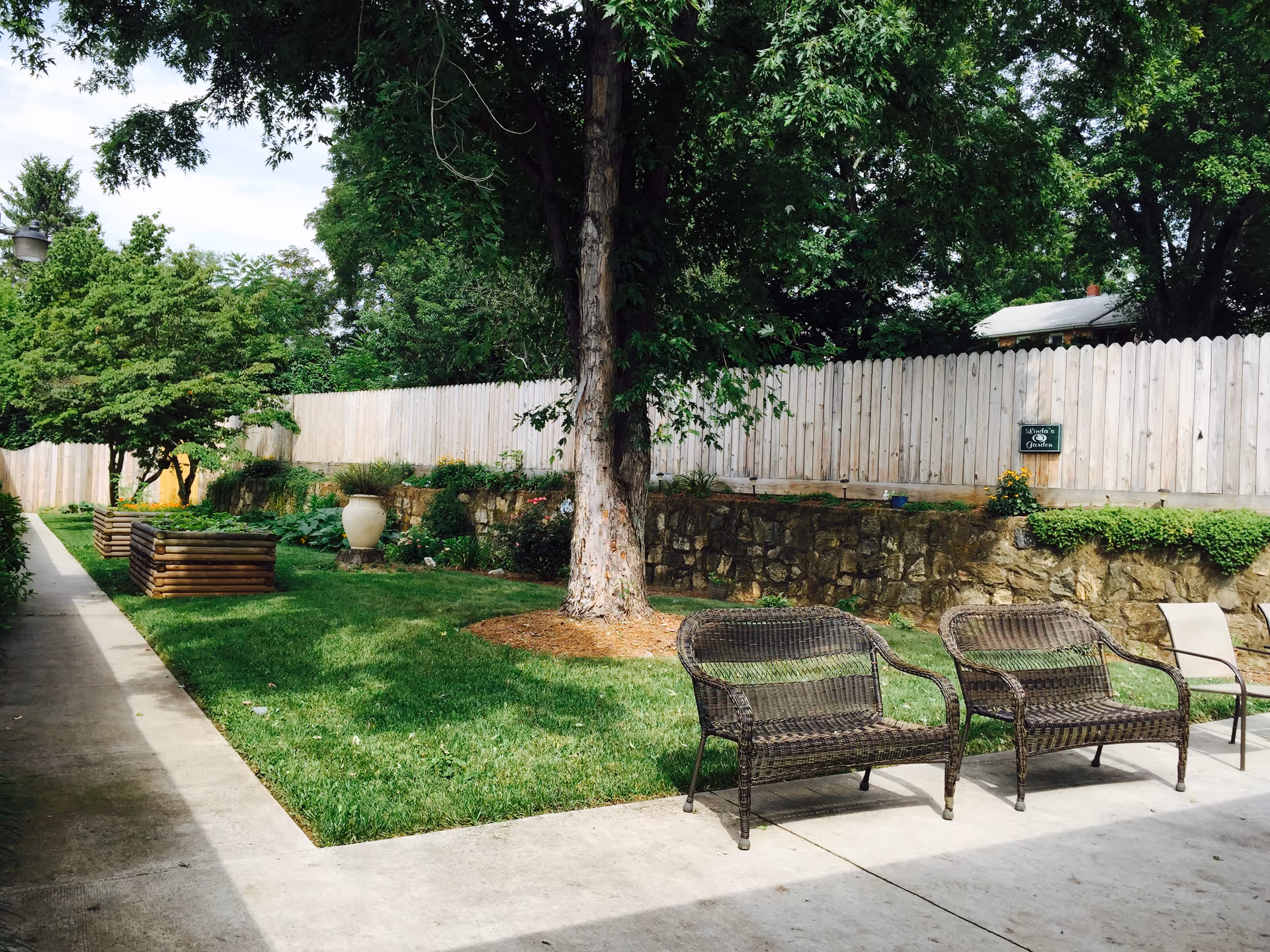 Outdoor garden area with green grass, a large tree, wooden raised garden beds, and a stone retaining wall topped with a wooden fence. There are two wicker chairs and one metal chair on a concrete patio. The area is shaded by large trees.