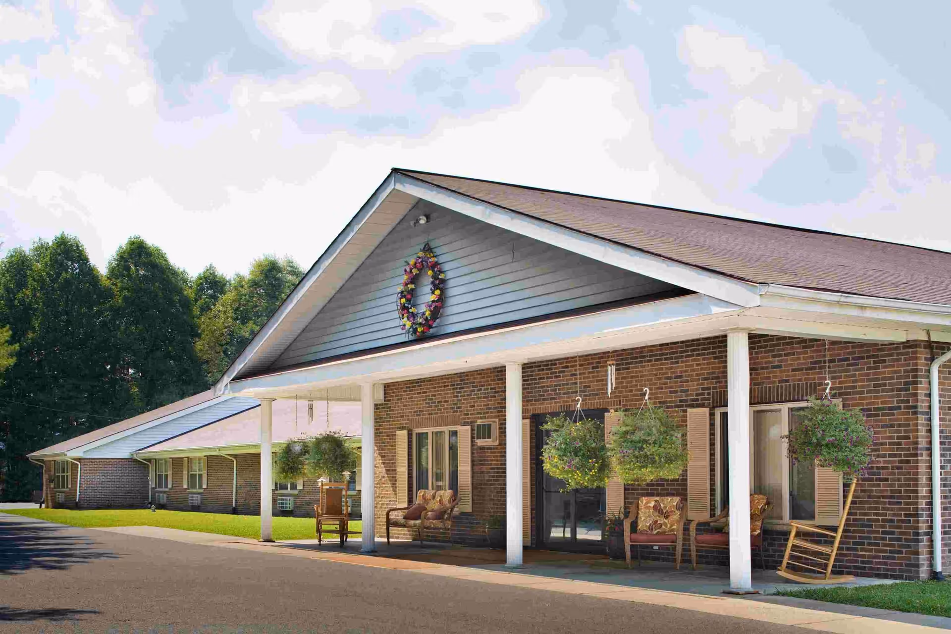 Exterior view of a single-story brick building with a covered porch supported by white columns. The porch has hanging plants, rocking chairs, and cushioned chairs. A decorative wreath is mounted on the front gable. Trees and a clear sky are visible in the background.
