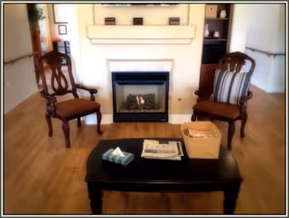 A cozy interior room featuring two wooden armchairs with cushions facing a black coffee table. The table holds a tissue box, newspapers, and a basket. Behind the chairs is a fireplace with a lit fire, flanked by built-in shelves and a window on the left side.