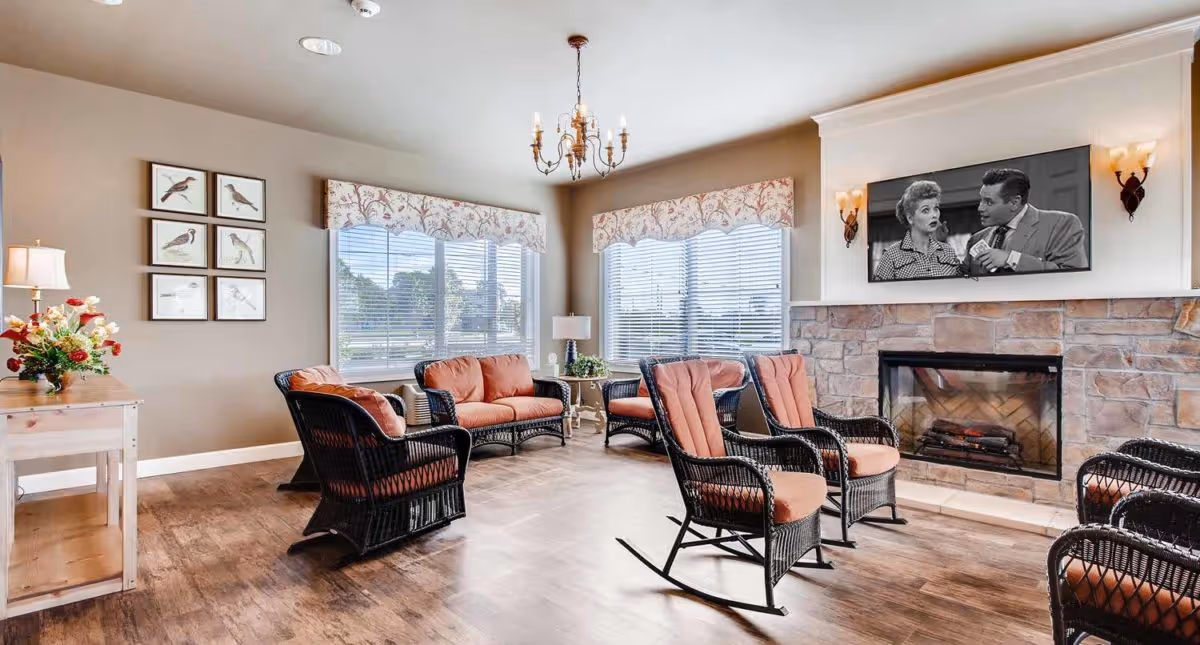 Bright communal living room with wicker chairs and cushions arranged around a fireplace and wall-mounted television.
