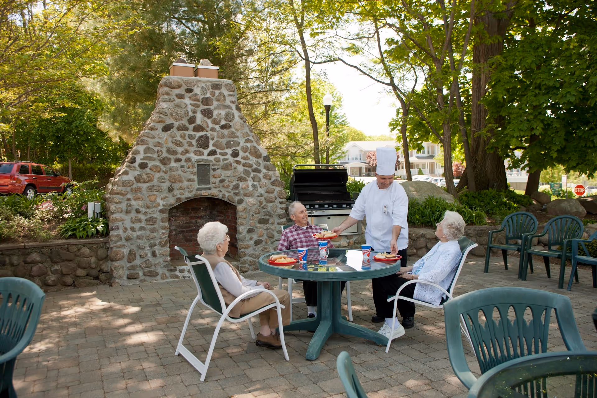 Three elderly people sitting around a green outdoor table with red plates and drinks, while a chef in white uniform serves food. They are in a patio area with a stone fireplace and a grill, surrounded by trees and greenery.