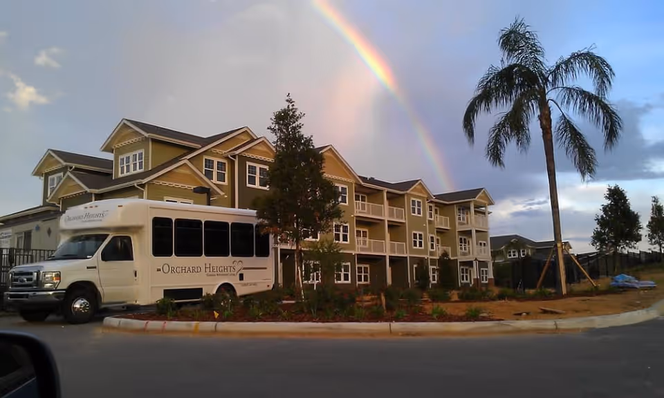 Exterior view of the Orchard Heights retirement building with a shuttle bus parked in front and a rainbow overhead.