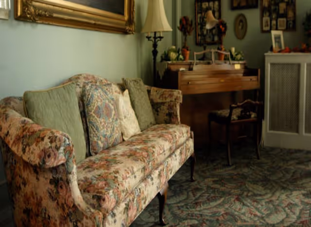 A cozy living room with a floral patterned sofa adorned with several cushions, a wooden piano against the wall with a chair in front of it, a tall floor lamp, and framed pictures hanging on the wall. The room has a carpet with a leafy pattern and a radiator cover on the right side.