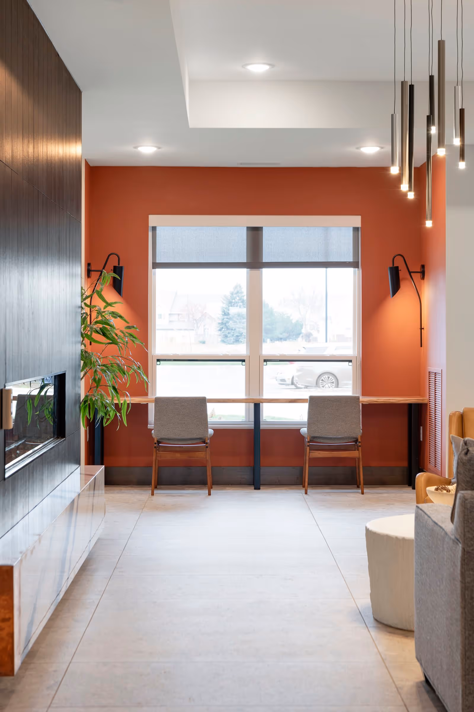 A modern interior space with a large window featuring a wooden desk and two gray chairs facing outside. The walls are painted a warm orange color, and there are two black wall-mounted lamps on either side of the window. To the left, there is a dark wood-paneled wall with a built-in fireplace and a green plant. On the right side, part of a gray sofa and a white round ottoman are visible. Several pendant lights hang from the ceiling on the right side.