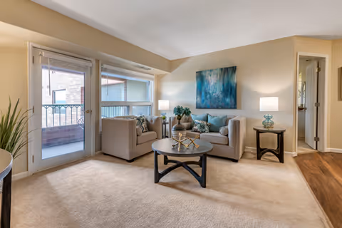 A cozy living room with beige carpet and walls, featuring two beige armchairs and a beige loveseat with blue and patterned cushions. A round black coffee table with decorative items sits in the center. Two side tables with lamps flank the loveseat. A large window and glass door lead to a balcony with a brick railing. A blue abstract painting hangs on the wall above the loveseat.