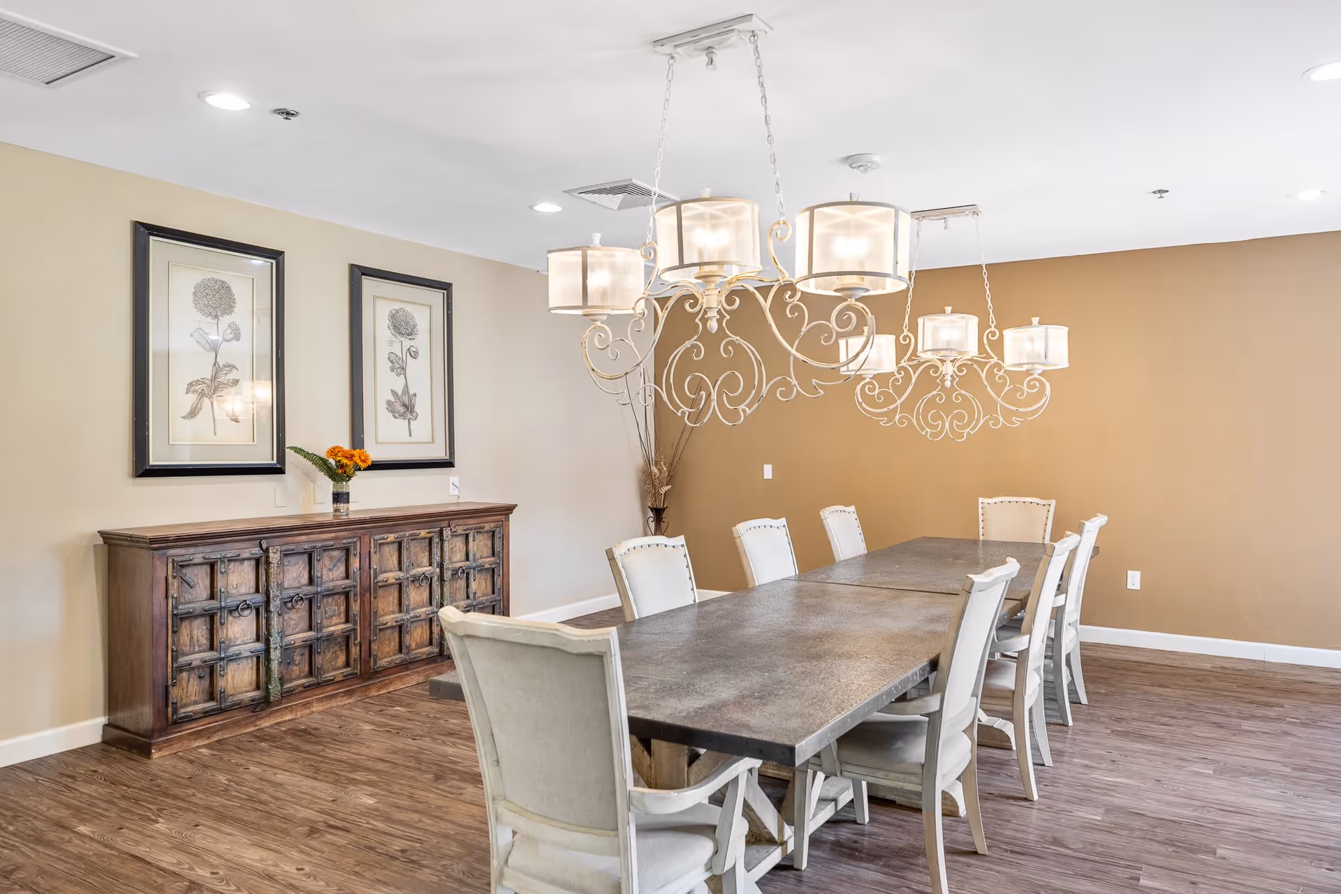 A dining room with a long rectangular table surrounded by eight white cushioned chairs. Two ornate chandeliers with multiple lights hang above the table. Against one wall is a wooden sideboard with a vase of orange flowers and two framed botanical prints hanging above it. The room has wood flooring and beige walls.