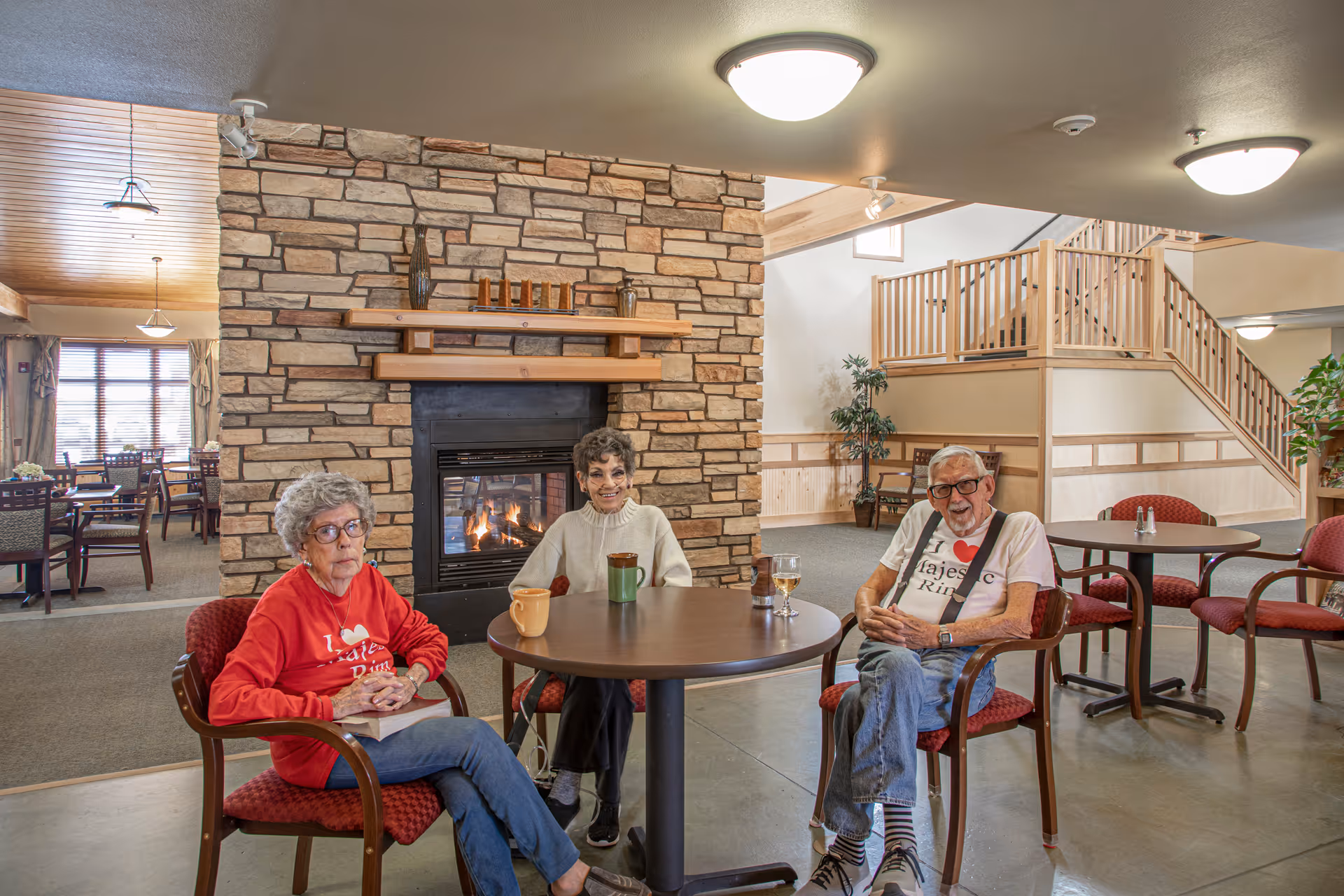 Three elderly people sitting around a round table in a cozy common area with a stone fireplace behind them. Two women and one man are seated on red cushioned chairs, with mugs and a glass on the table. The room has warm lighting, wooden accents, and a staircase in the background.