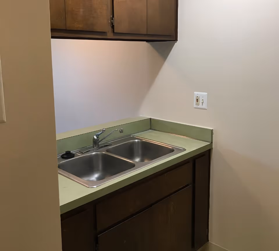 Small kitchenette with a double stainless steel sink, green countertop, and wooden cabinets.