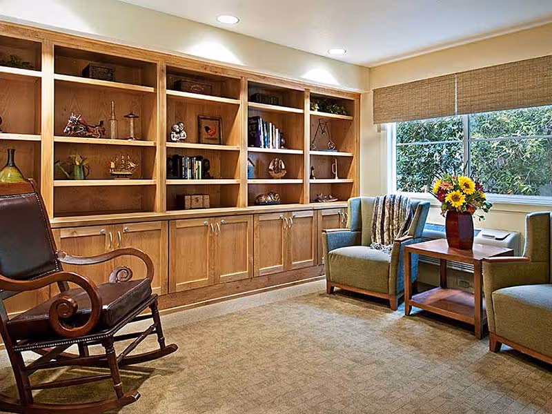 A cozy living room area with wooden built-in shelves filled with decorative items and books, a wooden rocking chair with dark leather upholstery, two green upholstered armchairs, a wooden side table with a vase of sunflowers, and a large window with beige blinds letting in natural light.