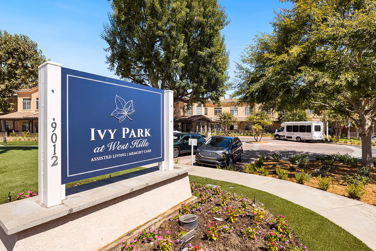 Entrance sign for Ivy Park at West Hills assisted living and memory care facility with a landscaped garden, parked cars, and the building in the background under a clear blue sky.