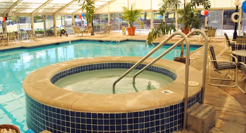 Indoor swimming pool area with a circular hot tub in the foreground surrounded by blue tiles. The pool area is enclosed with large windows and a glass ceiling, allowing natural light to fill the space. There are chairs and potted plants around the pool, and some red, white, and blue balloons are visible in the background.