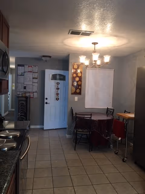Interior view of a kitchen and dining area with tiled floor. On the left, there is a kitchen counter with a stove and microwave. At the back, a white door with heart-shaped decorations is visible. A round dining table with a dark tablecloth and four chairs is positioned under a chandelier light fixture. A window with closed blinds and a decorative wall hanging are on the wall behind the table. A small cart with a red towel is on the right side.