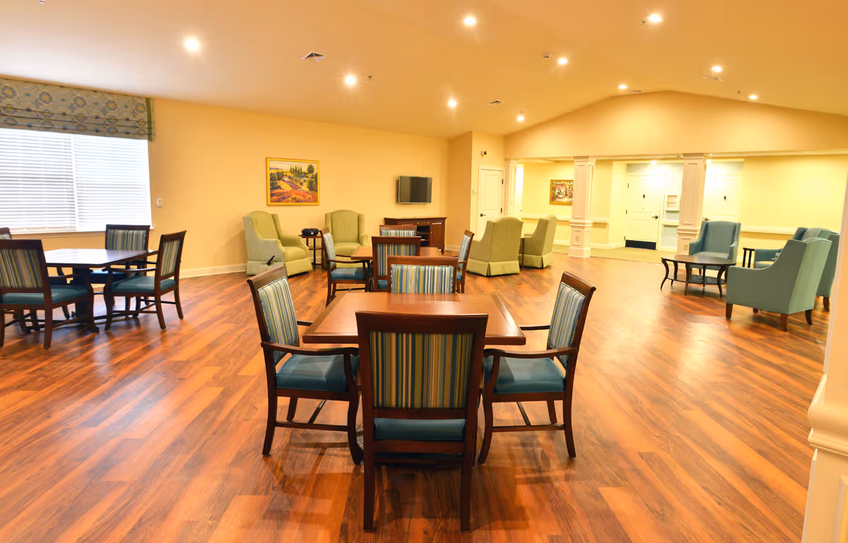 A spacious assisted living common area with wooden floors, several tables with chairs featuring striped upholstery, and multiple armchairs arranged in small seating groups. The walls are painted light yellow, and there is a window with blinds and a patterned valance. A flat-screen TV is mounted on the wall above a wooden cabinet, and framed artwork is displayed on the walls. The ceiling has recessed lighting.