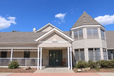 Front exterior view of a single-story building with beige siding, a peaked roof, and a turret-like structure on the right side. The entrance has a covered porch with white pillars and double green doors. The building number 1820 is displayed above the entrance. There are shrubs and rocks in front of the building under a blue sky with some clouds.