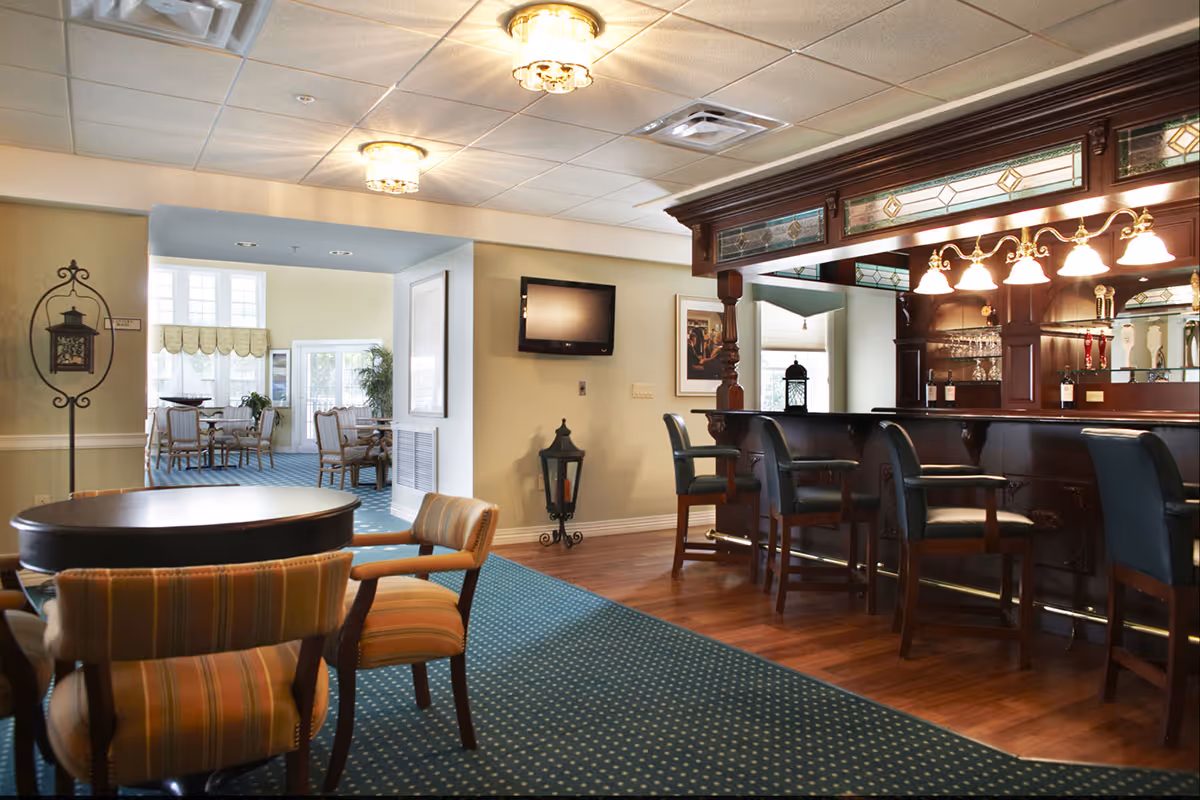 Interior view of a senior living facility lounge area featuring a wooden bar with high chairs on the right, a round table with striped upholstered chairs in the foreground, and a dining area with tables and chairs visible through an open doorway. The room has a green carpet with a dotted pattern, wood flooring near the bar, and ceiling lights.