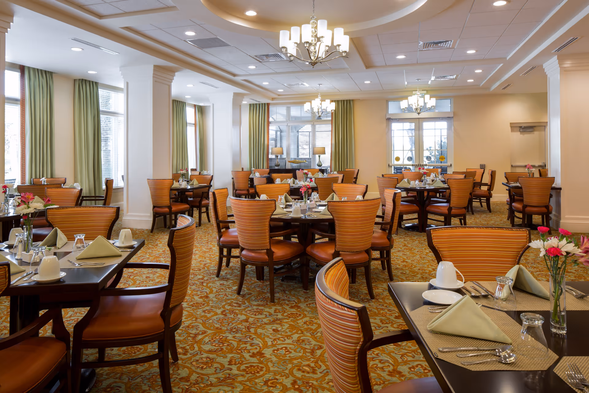 A spacious dining room with multiple round and rectangular tables set with green napkins, white cups, silverware, and small flower vases. The room features patterned carpet, large windows with green curtains, white walls with columns, and elegant chandeliers hanging from the ceiling.