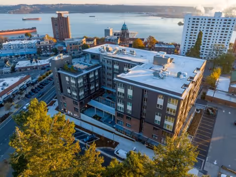 Aerial view of a multi-story senior living facility building with a flat roof, surrounded by trees and adjacent parking lots. The building is located near a waterfront with boats visible in the water and a cityscape in the background under a clear sky.